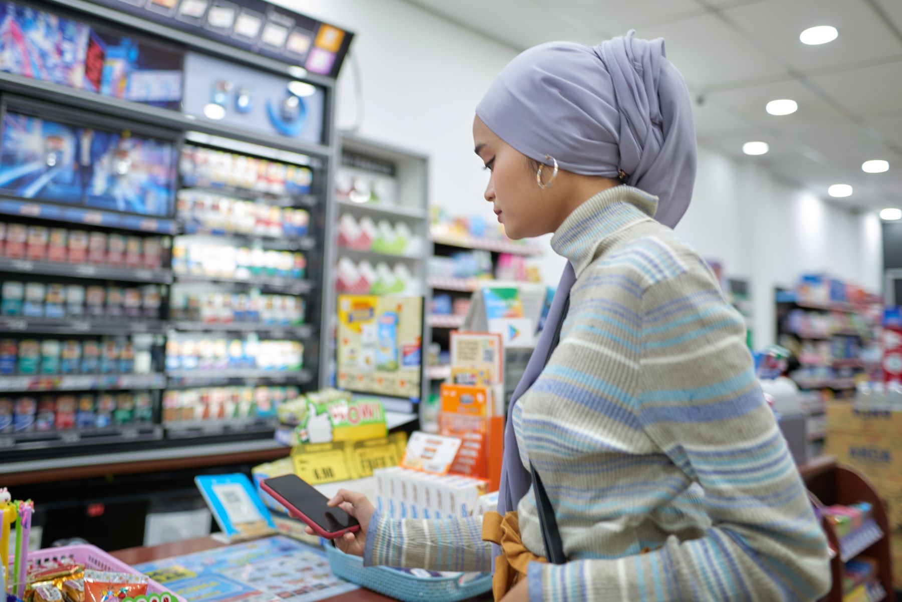 Woman in a headscarf and striped sweater using a smartphone inside a convenience store.